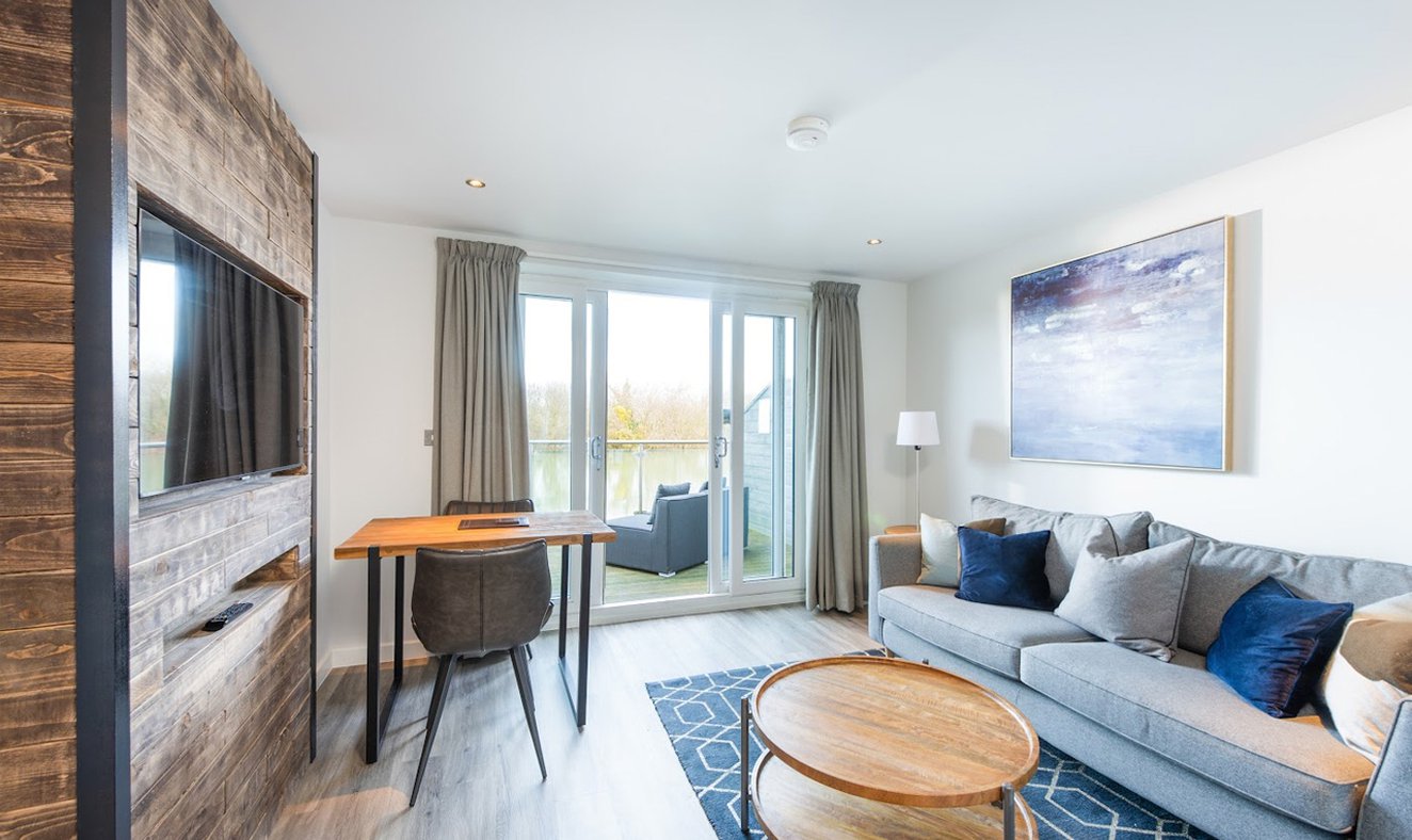 A living and dining area of one of our apartments, contains a three-seater sofa, a round coffee table, a TV mounted into the wall and a small dining table. In the background is a set of french doors leading to a balcony area.