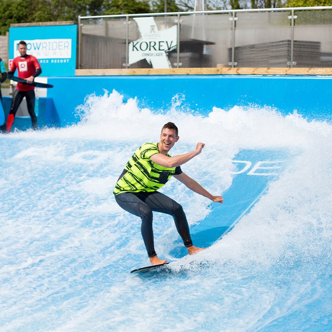 
                A young male is surfing on a wave stimulator with two other males watching from the sideline.
                