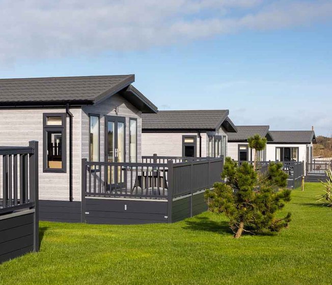 Some of our luxury lodges at one of our holiday parks. The view is of the exterior of the lodges, with the surrounding green space, and a small tree.