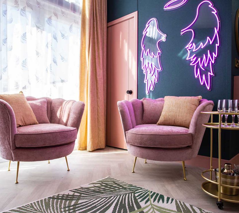 The stunning lounge area in The Esme Compact. There are two plush velvet chairs, a gold drinks trolley and some pink neon lighting in the shape of angel wings.