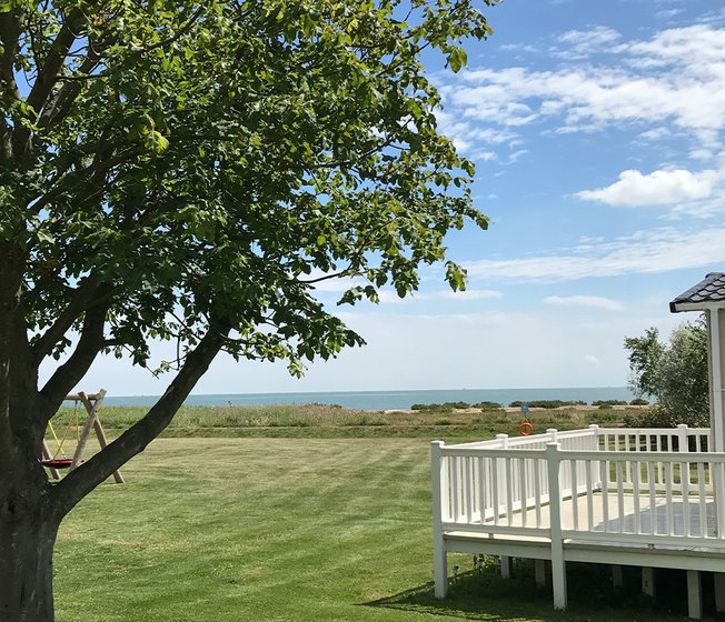 The grounds at our Mersea Island holiday park. There is a view of the porch of one of your lodges, a tall tree, a playground area, and plenty of outdoor green space.