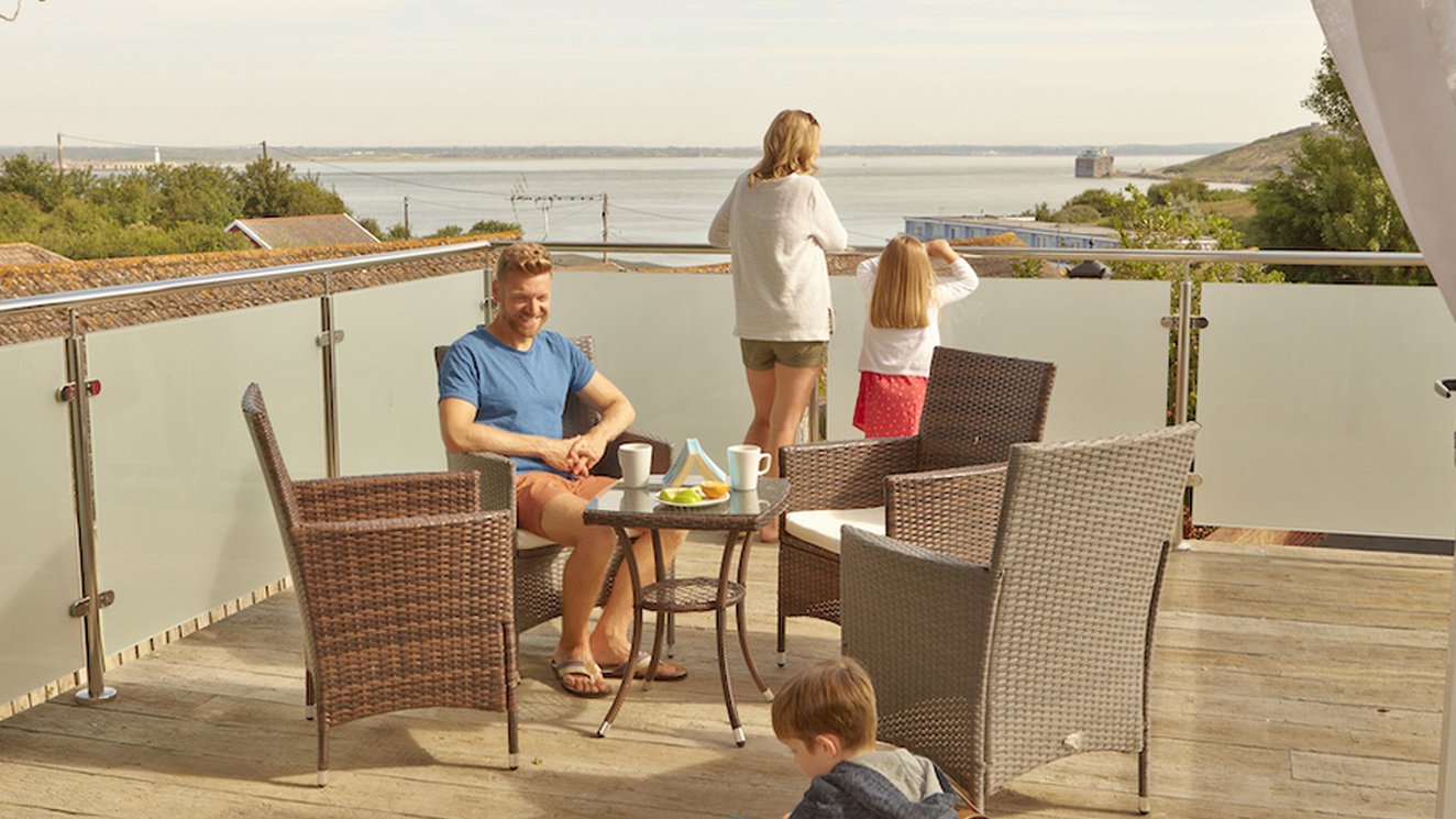 
              A family of four on the balcony of their holiday home in The Bay Colwell. There are two adults, a young boy, and a young girl with strawberry blonde hair, and all dressed in summer attire.
              