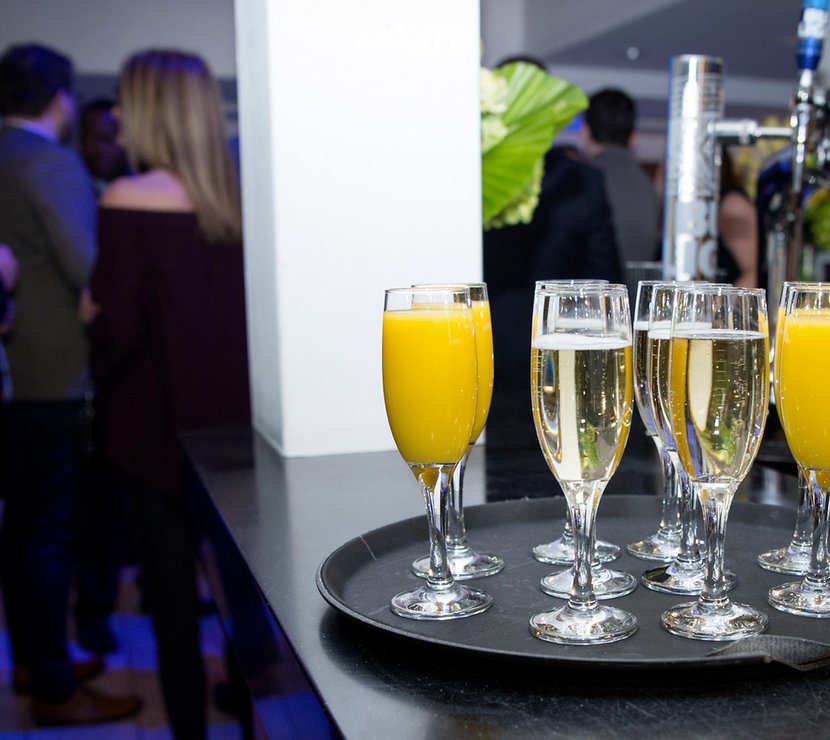 A black tray holds a selection of drinks in champagne glasses, either filled with fizz or orange juice, whilst a party occurs in the background with a group of people seen standing.
