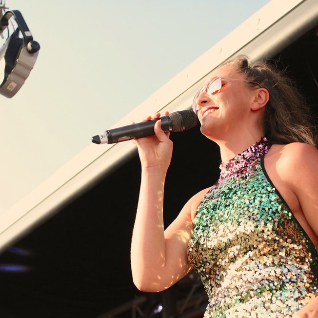 
                A woman with brown hair in a ponytail wearing a green, gold and purple sequin dress signing into the microphone.
                