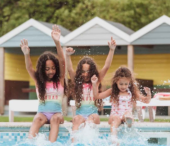 Three girls, each with long brown curly hair and pink swimsuits, are splashing and paddaling on the edge of our outdoor pool at Newquay Bay, Cornwall