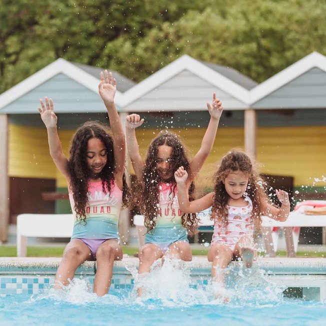 
                Three girls, each with long brown curly hair and pink swimsuits, are splashing and paddaling on the edge of our outdoor pool at Newquay Bay, Cornwall
                