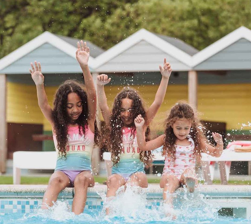 Three girls, each with long brown curly hair and pink swimsuits, are splashing and paddaling on the edge of our outdoor pool at Newquay Bay, Cornwall