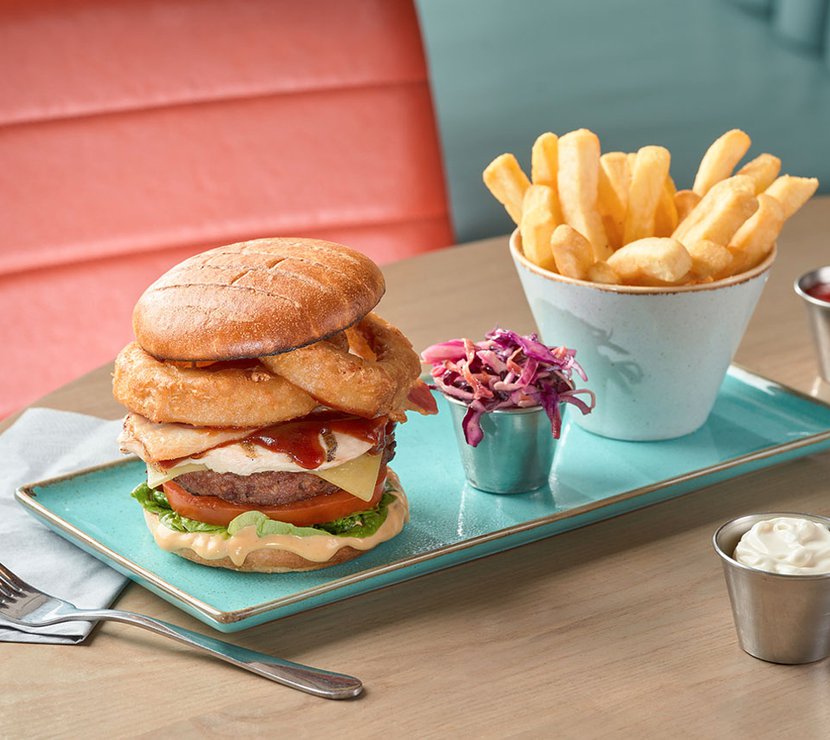 A rectangle plate with a white ceramic holder of chips, a tall stacked burger and a small metail container with red cabbage.