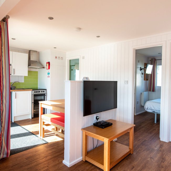 
                The interior of one of our holiday home Chalets. There’s a large TV, double bedroom, and kitchen space. There are bright colours in the holiday home.
                