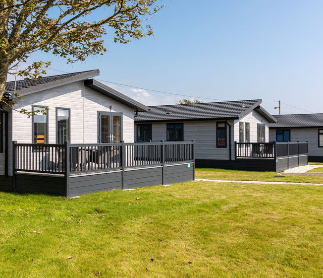 Our amazing lodges at one of our holiday parks. There are three luxury lodges surrounded by green grass, and the sun is shining down.