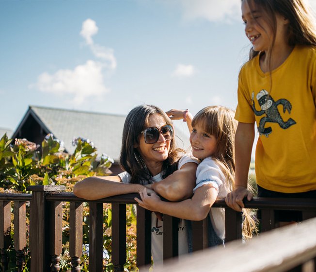 A family enjoying their stay at one of our holiday parks. There is an adult woman and two young girls. One of the girls is hugging the woman, the other is leaning over a banister. All three have wide smiles.