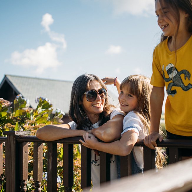 
                A family enjoying their stay at one of our holiday parks. There is an adult woman and two young girls. One of the girls is hugging the woman, the other is leaning over a banister. All three have wide smiles.
                