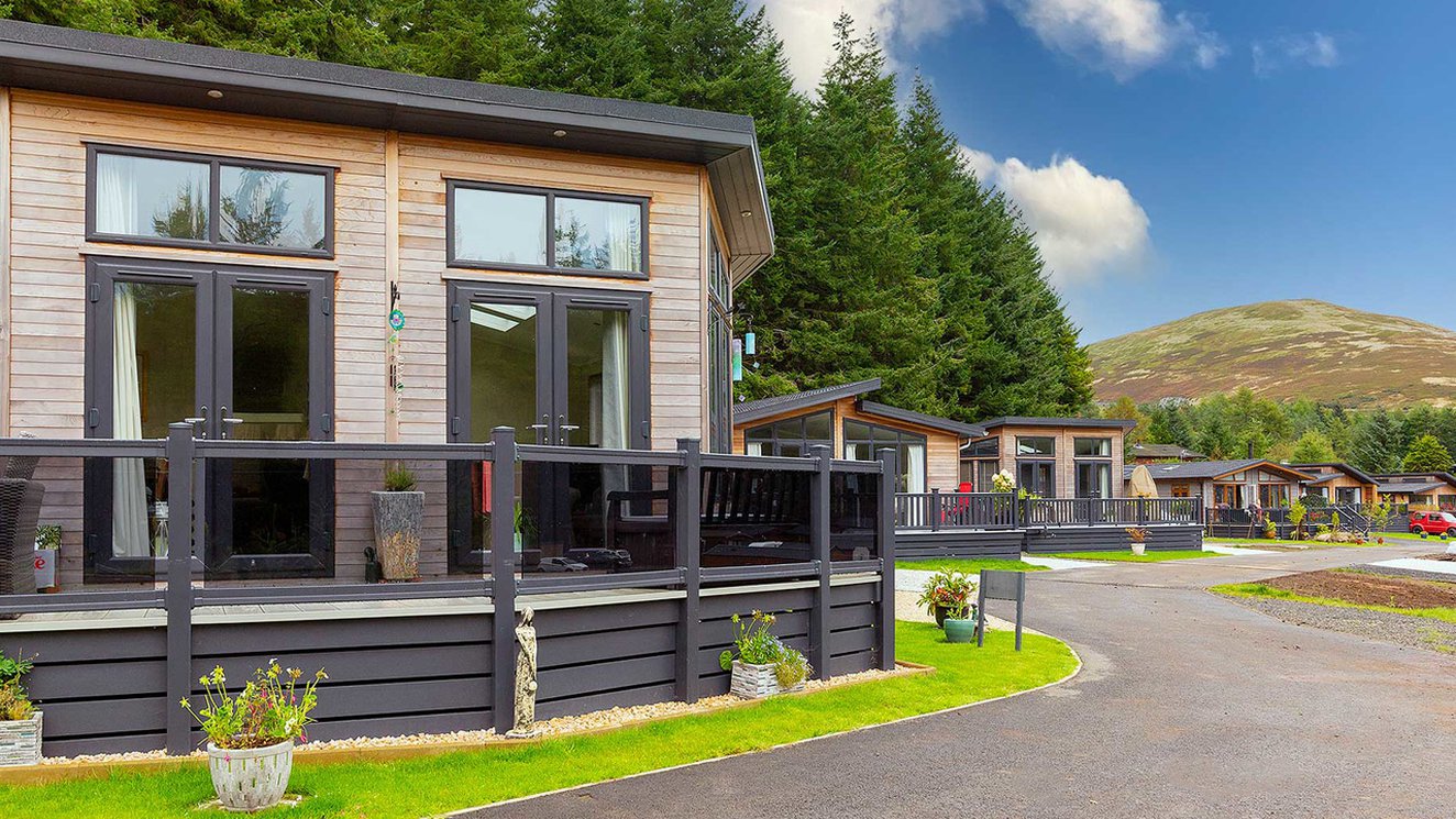 
              A view of the exterior of our stunning holiday homes for sale at Glendevon Residential Country Park, Scotland. There is a row of wooden lodges and views in the background of the stunning surrounding scenery.
              