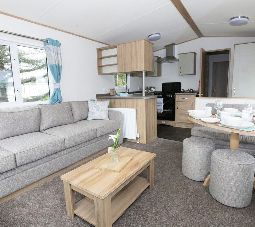Suggested: The interior of one of our luxury caravans. There's a large, grey sofa, a seating area with a table, and a kitchen space with several appliances.