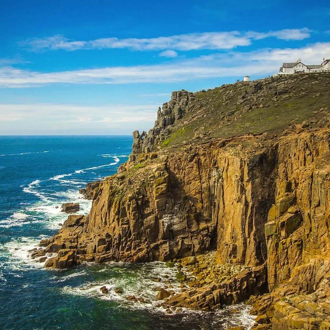 
                A landscape image of the sea and cliffs located at Lands End. A blue sky with minimal clouds sets the scene with the Land's end tourist building seen in the top right of the image.
                