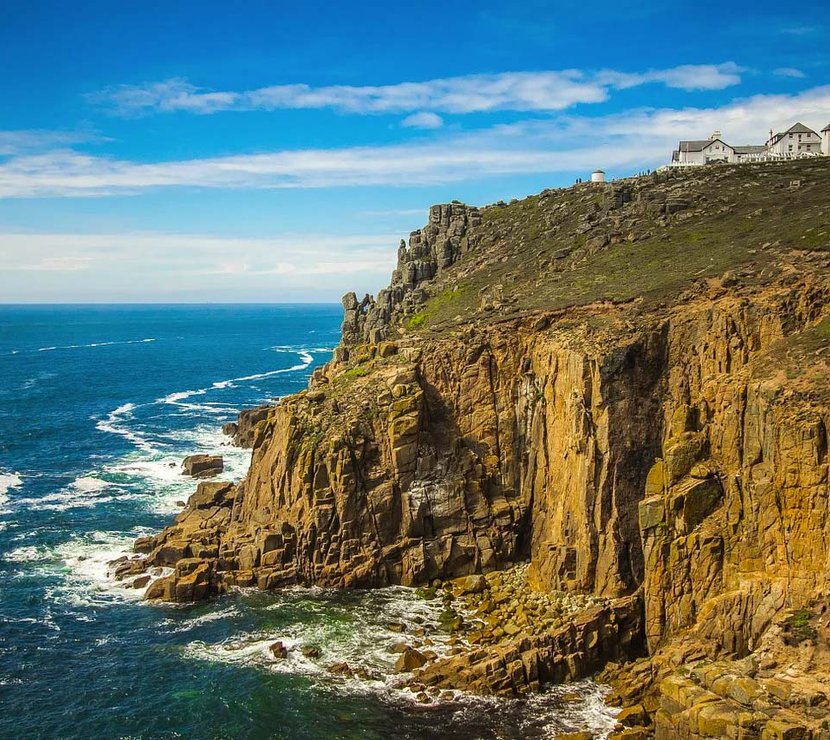 A landscape image of the sea and cliffs located at Lands End. A blue sky with minimal clouds sets the scene with the Land's end tourist building seen in the top right of the image.