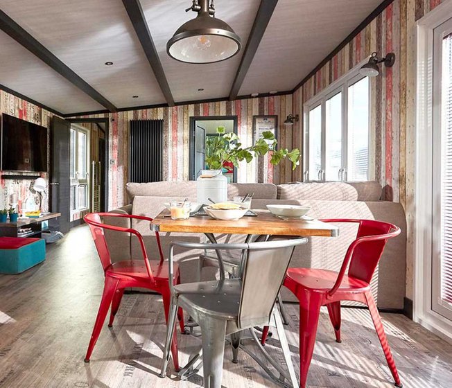 An image of the interior of one of our holiday lodges. There's wooden panneling, a cream fabric sofa and red metal dining chairs around a wooden table.