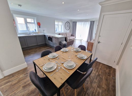 An open-plan view of one of our cottages. In front view is a dinner table set for six. across from the table is a U-shaped Kitchen area with plenty of worktop space. In the background is the living room area containing two sofas.
