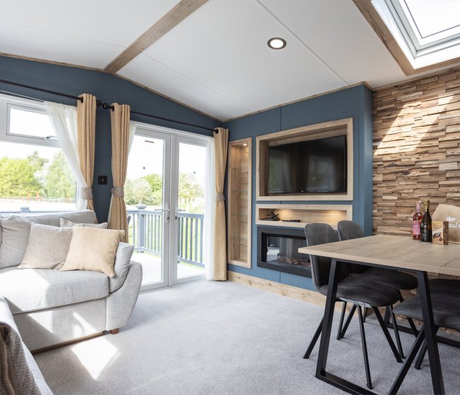 The inside of one of our static accessible caravans. There is a grey sofa and a dining area, along with a TV and fireplace. The large double doors are letting in sunlight.
