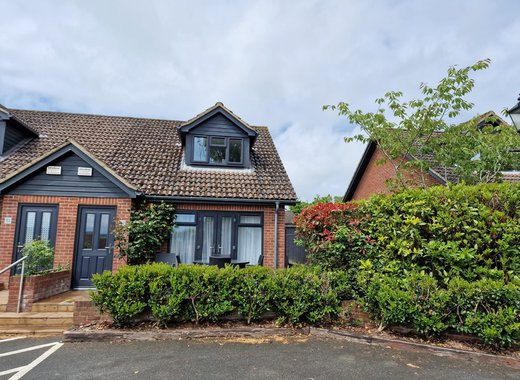 An outside view of one of our 3-bed cottages. Bushes outside lead to three steps to the pathway of the front door to the cottage. The sky above is blue yet very cloudy.