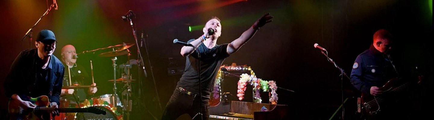 
          An adult male on a stage singing at a rock gig. He's got short light brown hair, a black tee and dark jeans singing into the microphone and pointing out to the crowd.
          