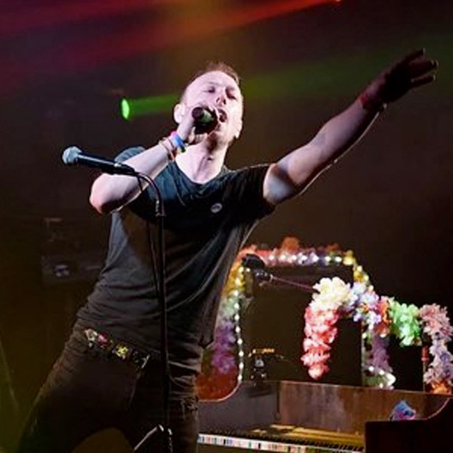 
                An adult male on a stage singing at a rock gig. He's got short light brown hair, a black tee and dark jeans singing into the microphone and pointing out to the crowd.
                