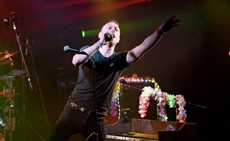 An adult male on a stage singing at a rock gig. He's got short light brown hair, a black tee and dark jeans singing into the microphone and pointing out to the crowd.
