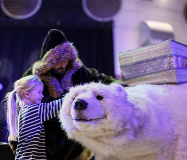 A man wearing a black hooded cloak with silver trim is petting a white life-sized plush of a polar bear with a little girl with blonde hair in a ponytail and a striped long sleeved t shirt.