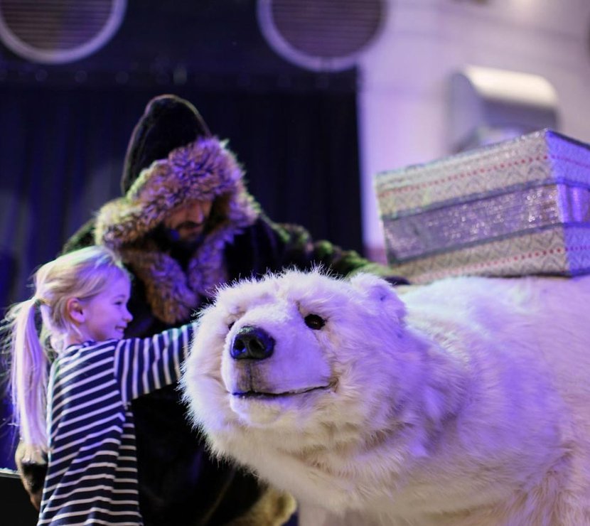 A man wearing a black hooded cloak with silver trim is petting a white life-sized plush of a polar bear with a little girl with blonde hair in a ponytail and a striped long sleeved t shirt.