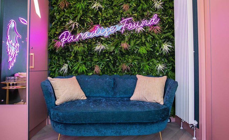 A uniquely decorated seating area with fake vines on the wall, a blue loveseat and a pink neon sign which says, ''Paradise Found".