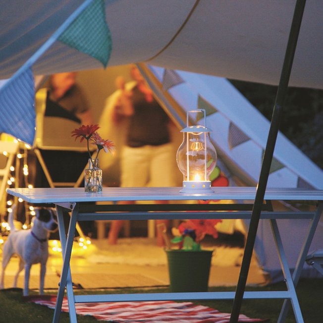 
                A sun set evening with a Jack Russell dog standing at the entrance of a off-white canvas bell tent. There is also a blue and white picnic table next to the dog.
                