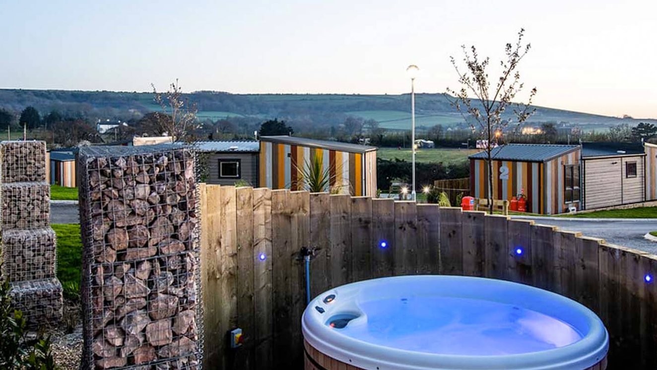 
              The outside of one of our holiday parks, which has an empty hot-tub behind a private fence. The sun is setting behind the wide view of the holiday park.
              
