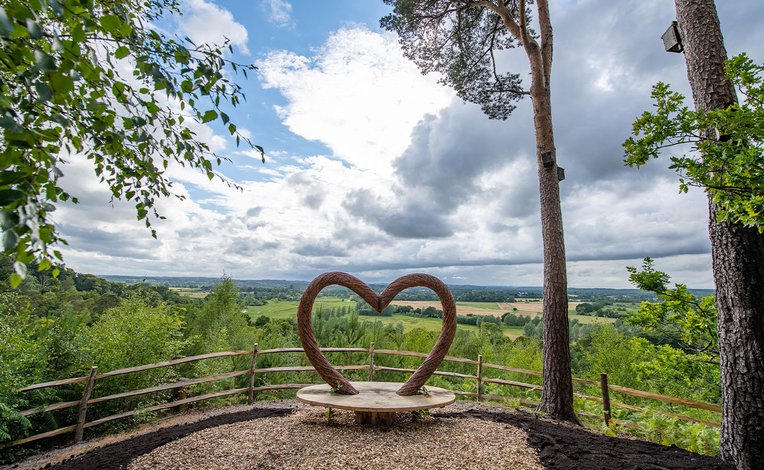 A panoramic view of the countryside, with a viewing area in the foreground. The bench features a wooden heart arch.