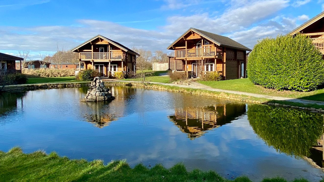 
              Two of our stunning lodges at Woodland Lakes, overlooking onto a small lake on a sunny, cloudy day with blue skies.
              