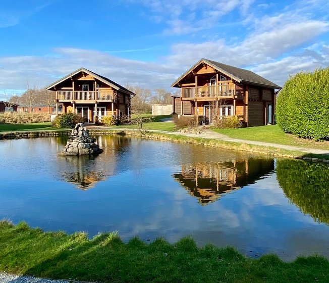 Two of our stunning lodges at Woodland Lakes, overlooking onto a small lake on a sunny, cloudy day with blue skies.