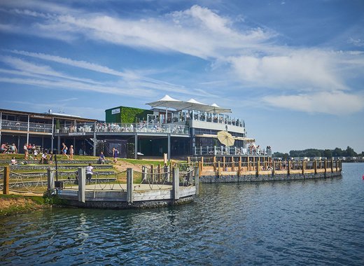 A photo of a restaraunt with a ground floor and two balconey levels of outdoor steating. There are lots of people sitting across all levels and the building is next to a large river.