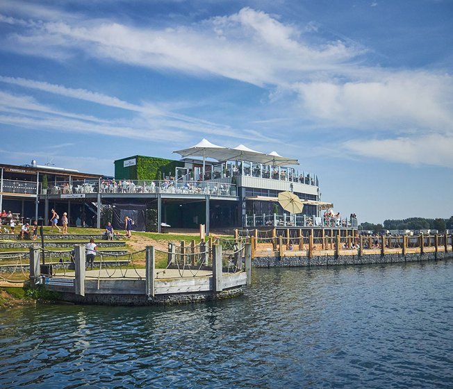 A photo of a restaraunt with a ground floor and two balconey levels of outdoor steating. There are lots of people sitting across all levels and the building is next to a large river.
