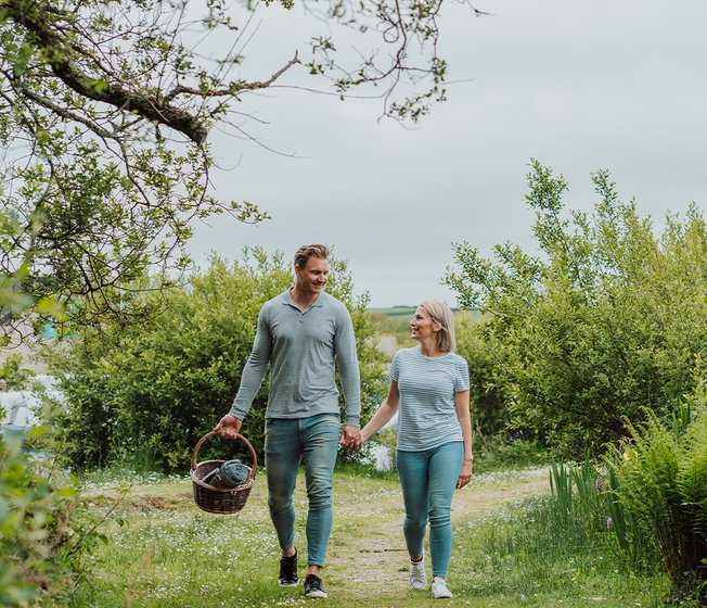 A young white couple walk hand in hand down a country path whilst the male holds a picnic basket.