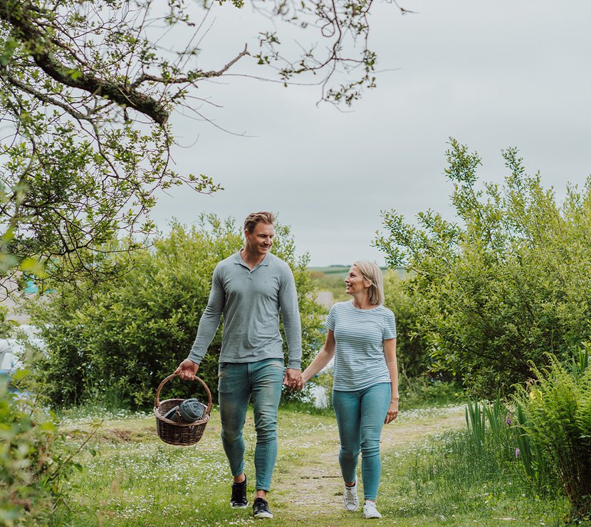 A young white couple walk hand in hand down a country path whilst the male holds a picnic basket.