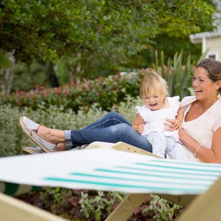 An adult white woman and young child are outside sitting on a wooden deck chair. The child is sat on the lap of the woman and has a gleeful expression. The woman is suporting the child upright and is smiling.