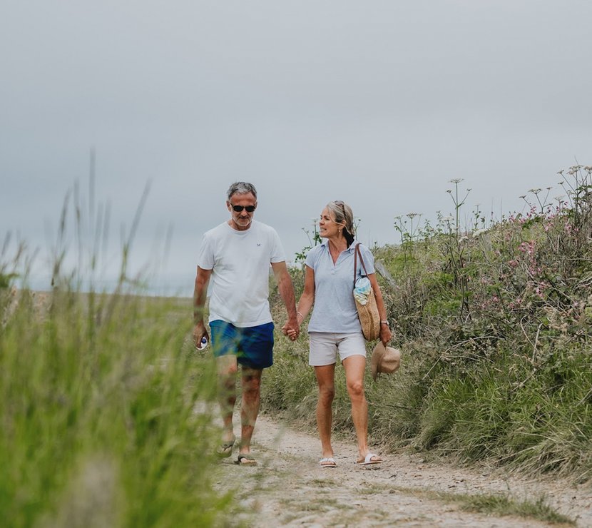 A white, middle-aged couple walking hand in hand down a country path. The man is wearing a white t-shirt with blue shorts. The woman is wearing a light blue polo shirt with cream shorts, whilst holding a weaved hat in her hand.