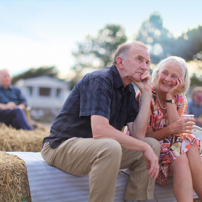 
                An older couple sat on a hay bale on a sunny day.
                