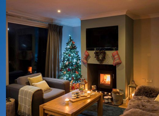 An interior photo of a decorated chimney brest with a lit wood burner. On the wall above it there is a heart-shaped reath, two red stockings either side and on the floor there is a bucked of logs and a pair of boot slippers.
