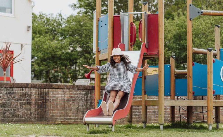 A wooden children's adventure playground outside on some trimmed grass at Newquay Bay, Cornwall. A girl wearing a white hat and a checkered dress is sliding down a red slide.