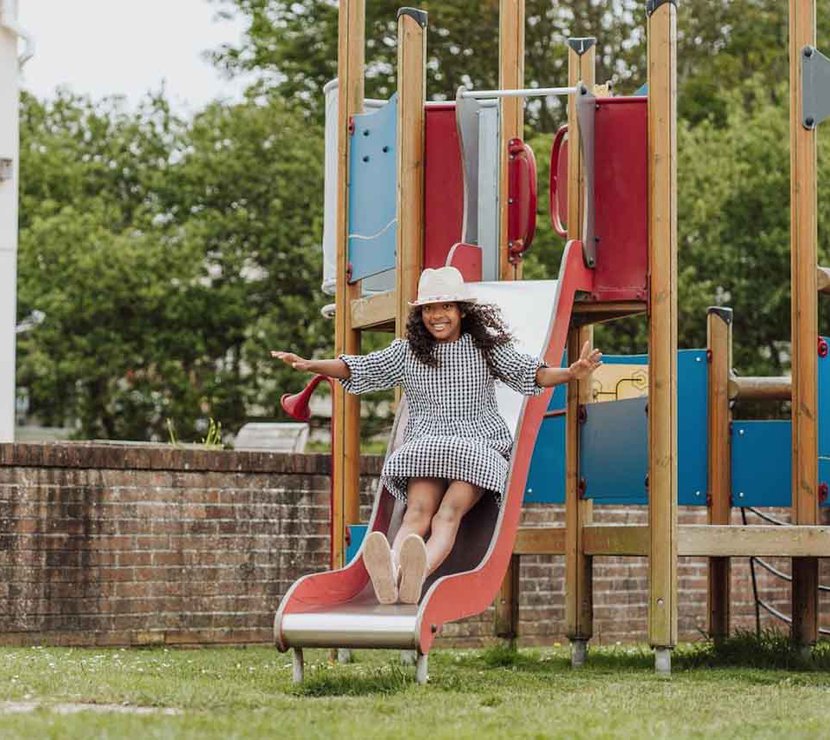 A wooden children's adventure playground outside on some trimmed grass at Newquay Bay, Cornwall. A girl wearing a white hat and a checkered dress is sliding down a red slide.