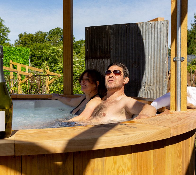 A middle-aged couple sitting in a bubbling hot tub under a canopy. On the edge of the hot tub is a bottle of prosecco.
