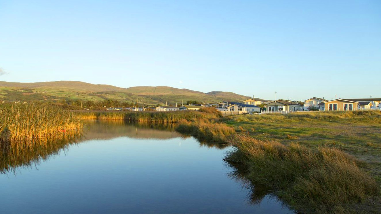 
              A view of our holiday park in North Wales, Barmouth Bay, in the early evening. There is a view of the lake, a row of holiday homes and mountains in the distance.
              