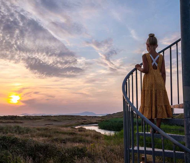 A photo of a girl with blonde hair in a bun wearing a orange polkadot dress, whom is standing in the middle of a spiral staircase looking out at the sunset.