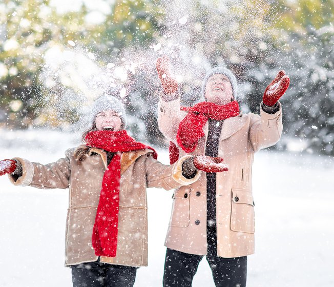 A man and a woman in light brown winter coats, red gloves and scarfs and grey benie hats are throwing snow up in the air and laughing as the snow crashes down on them.