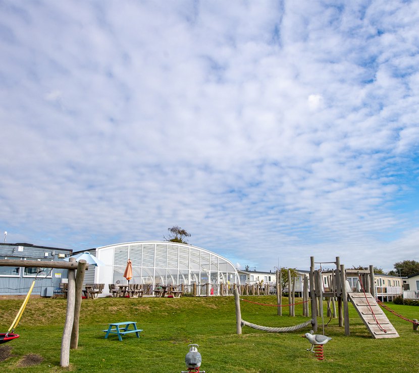 The outdoor adventure playground at our Essex holiday park, Mersea Island. There is views of the indoor swimming pool conservatory and some caravans in the background.